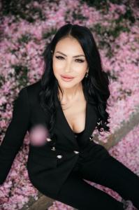 Giulia Deamina seated among pink blossoms, wearing a fitted black blazer, with long black hair and looking toward the camera.