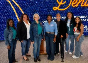 Photo with seven people smiling in front of a screen with a digital denim background. See caption for names and titles.