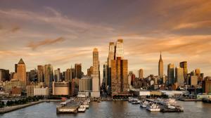 A golden hour view of the New York City skyline featuring the Empire State Building and other skyscrapers, with several white ferries docked at a Hudson River pier in the foreground.