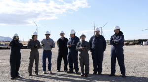 A group of professionals wearing white hard hats and protective workwear conducting an EB-5 project site visit at the Utah Cove Fort Geothermal Plant, with wind turbines visible in the background.