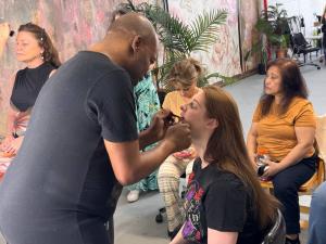 A professional makeup artist applies lip color to a young woman with long red hair during a VIP Ignite Certified Production Assistant Program hands-on training session, with additional participants and instructors visible in the background of a studio env