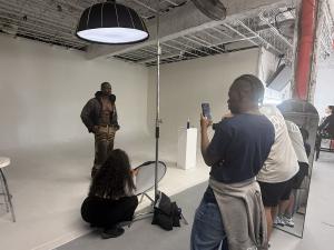 VIP Ignite Certified Production Assistant Program participants observe and assist during a live professional studio photo shoot, with a male model posed against a white seamless backdrop while trainees manage lighting equipment and camera positioning in a