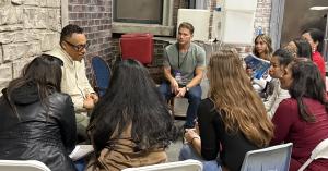A VIP Ignite industry professional wearing a talent credential badge leads an intimate small-group mentoring session with aspiring models and actors seated in a circle inside a Hollywood studio set with exposed brick walls, providing personalized career g
