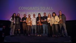 group of 10 filmmakers standing on stage holding their awards that they just won and smiling while a background on the planetarium screen behind them says congrats