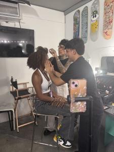 Two professional makeup artists apply makeup and style the hair of a VIP Ignite talent seated in a director's chair during a live Hollywood event, with a fully equipped professional makeup station visible in the background, preparing the participant for p