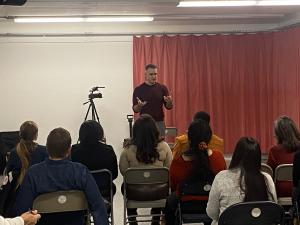 A working entertainment industry professional stands at the front of a room addressing a diverse group of aspiring models and actors seated in rows during a VIP Ignite industry networking event, with a professional video camera on a tripod capturing the s