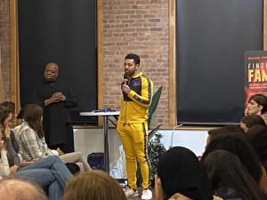 A working entertainment professional addresses a packed room of aspiring models and actors at a VIP Ignite industry networking event in New York, speaking into a microphone while an audience listens attentively, with Sir Michael Fomkin's book Finding Fame