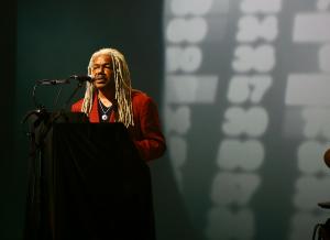 Landry Butler wears a red suit and stands in front of a screen full of numbers during Inglewood Social Club's performance of Welcome to Paradise.