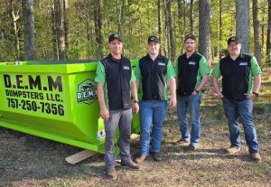 Four Navy veteran founders of D.E.M.M. Dumpsters LLC standing next to a green roll-off dumpster in matching branded uniforms.