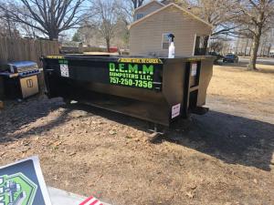 Black D.E.M.M. Dumpsters roll-off delivered in a residential backyard with bilingual safety labeling and visible phone number.