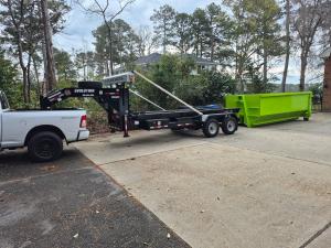 White Ram pickup with gooseneck trailer staging a green D.E.M.M. Dumpsters roll-off for residential delivery in Hampton Roads.
