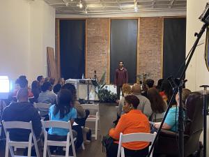 An aspiring actor stands before a packed room of industry professionals and fellow talent during a live performance showcase at a VIP Ignite industry networking event, with professional lighting and video equipment visible and a diverse audience seated in