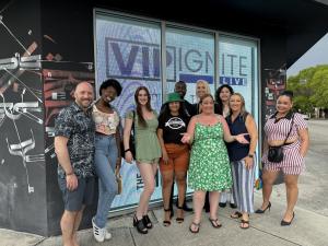 A diverse group of aspiring models and actors smile and pose together outside a VIP Ignite LIVE event venue in Miami, with the illuminated VIP Ignite LIVE logo displayed prominently in the window behind them, representing the energy and diversity of South