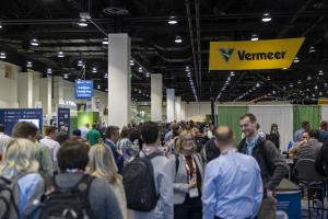 Crowded trade show floor with attendees networking in a large convention hall, featuring a prominent yellow Vermeer sign overhead and various exhibitor booths and banners in the background.