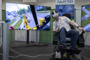 Person seated in a VR excavation simulator at a trade show booth, wearing a headset and operating joystick controls while a large screen displays a first-person view of heavy equipment digging along a residential street.