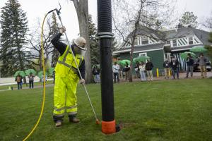 Worker in high-visibility gear uses a pressurized wand to excavate soil around a utility pole on a lawn while a group of onlookers stands nearby, many holding green umbrellas, in front of a residential building.