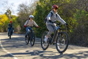 Three teenagers wearing bicycle helmets ride e-bikes along a paved path bordered by trees and greenery on a sunny day.