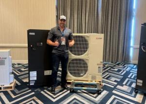 Christopher Hernandez, CEO of Arrow Appliance, giving a thumbs up while standing between a black HVAC unit and a white Mitsubishi outdoor condenser unit at an industry event.