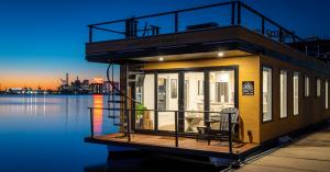 LOHOM 1 | Bay Escape, a two-story modern floating suite at Inner Harbor Marina in Baltimore, photographed at dusk. Warm light glows through floor-to-ceiling glass doors onto a wood deck with two lounge chairs, while a spiral staircase leads to the rooftop