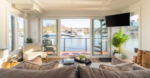 Interior of FLOHOM 1 | Bay Escape's living room featuring a gray sectional sofa with throw pillows, a cream accent chair, two round black coffee tables, and a wall-mounted TV. Floor-to-ceiling sliding glass doors open onto a wood deck overlooking Baltimor