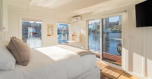 Bedroom inside FLOHOM 1 | Bay Escape with a queen bed dressed in white linens and a taupe throw, a wall-mounted TV, a fold-down desk, and a nautical chart on the wall. Floor-to-ceiling windows and sliding glass doors on three sides open onto views of Balt