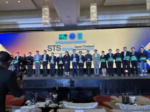 Roughly eighteen speakers standing in a line on stage holding green plaques and folders during the Appreciation Ceremony of STS forum Japan–Thailand Symposium 2026, with a large backdrop showing the STS forum, NRCT, and Thai government logos.