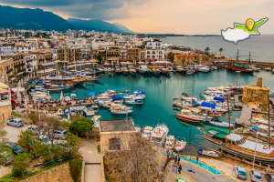 Aerial view of Kyrenia Harbour in Northern Cyprus showing colourful boats moored in turquoise water surrounded by historic waterfront buildings, restaurants, and the Kyrenia mountain range at sunset with NorthCyprus-Guide.com logo in top right