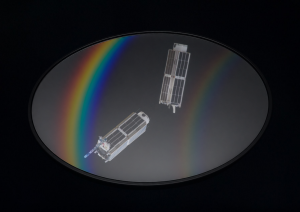 A shiny optic with curved rainbow spectra from it also reflects the images of CubeSats. The optic is over a dark background.