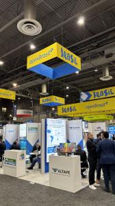 Indoor business trade fair environment with a high ceiling, exposed metal structure, and strong lighting. Suspended cubes in yellow and blue at the top mark the Brazil area. Below, there are organized booths with counters, panels, and monitors displaying