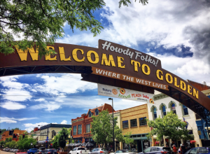 a brown wooden arch with lettering that reads Welcome to Golden! Where the West Lives spans a downtown street with green tree branches in foreground.