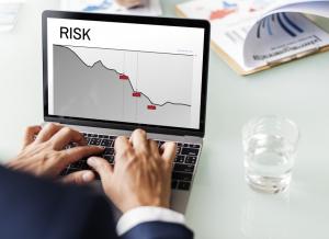 Businessman’s hands using a laptop displaying a risk analysis graph, with a glass of water and printed charts nearby