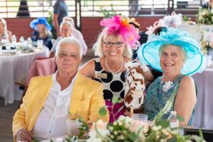Gary Sharpe, NTRC Executive Director Michelle Schweber, and Connie Sharpe smile together at a decorated Kentucky Derby Party table, wearing festive Derby-style hats and colorful attire while holding a plush stick horse.