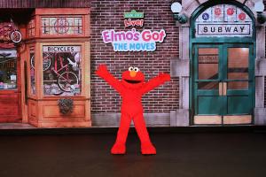 Elmo stands joyfully with outstretched arms in front of a brown brick backdrop featuring a bicycle shop and subway entrance, under "Elmo's Got the Moves" sign.