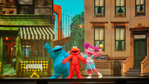 Three colorful, furry characters stand on a stage in front of a vibrant city backdrop. They exude joy, with arms raised in celebration.