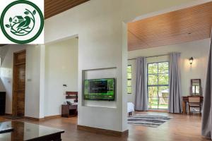 A photograph of a modern hotel suite interior with wood floors and a logo in the top-left corner. The central white room divider features an embedded television displaying a 'WELCOME' nature scene. To the right, a large window with grey curtains reveals t