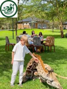 A photograph taken at Aya Sophia Wildlife Resort showing a young boy in white petting a giraffe resting on a grassy lawn. In the background, adults dine at a table with animal-print chairs, with a resort building visible behind them.