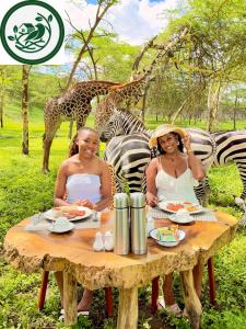 Two women smile while eating breakfast at a rustic wooden table outdoors at the Aya Sophia Wildlife Resort. Immediately behind them, giraffes and zebras roam freely across a lush, green landscape dotted with trees.