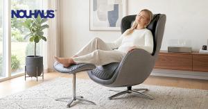 Woman relaxes with eyes closed in a grey and black quilted Nouhaus New Classic Massage Chair with feet on matching ottoman. Set in a bright, modern living room with large window, potted plant, and sleek media console. Blue NOUHAUS logo in upper left.