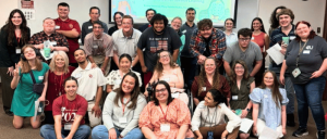 Group photo of diverse adults in a classroom setting, some seated and others standing, with a presentation screen in the background.