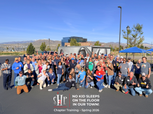 A large group of approximately 80 smiling volunteers give thumbs up as they pose together outdoors in a parking lot in Lehi, Utah, with snow-capped mountains visible in the background. An SHP branded trailer is visible behind the group. A graphic overlay 