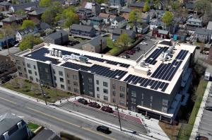 A new, large multifamily residential building sits on the corner of a busy street. The roof is covered in dark blue solar panels.