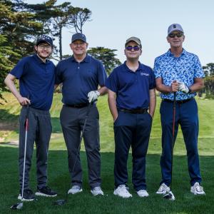 A photo of four men smiling while standing on the golf greens holding golf clubs and wearing blue golf attire.