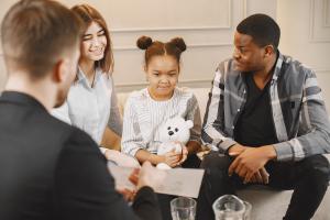 A young girl holding a white teddy bear sits between two adults during what appears to be a counseling or support session, with a professional visible in the foreground.