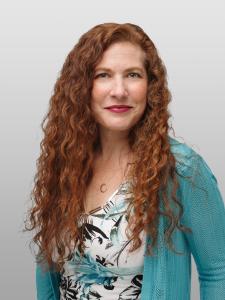 Janeen Marquardt, PMP, stands in a blue sweater with red wavy hair. She has recently earned the award of Distinguished Cheetah Alum for Significant Impact.