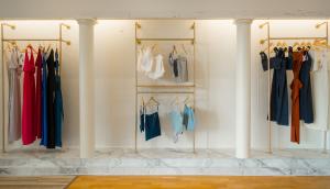 Interior of the Jendue premium denim store at Town Center Boca Raton, featuring three wall alcoves with gold clothing rails mounted between white columns on a marble floor. Left rail displays colorful ready-to-wear pieces including red, blue, black, and g