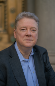 Professional portrait of veteran crisis communications advisor Tim Brown wearing a dark suit jacket and light blue shirt, seated indoors with a calm, confident expression against a softly blurred architectural background.