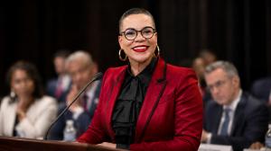 A Black woman in her mid-50s stands at a podium delivering a keynote presentation, dressed in a sharp blazer, expression commanding and composed.