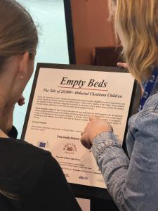 Two people viewed from behind reading an informational exhibition placard at the Berlaymont titled "Empty Beds: The Tale of 20,000+ Abducted Ukrainian Children.