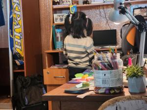 A person viewed from behind standing at a wooden desk inside a 1:1 recreation of a Ukrainian child's bedroom, examining the items left behind.