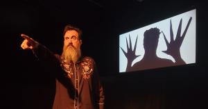 Comedian and filmmaker Jon Schnitzer with a long beard points to the left while a projection of a shadow man with wide hands is displayed on a screen behind him. Something Spooky is a paranormal comedy show that will give you goosebumps.
